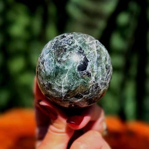 Puede incluir: Una esfera de cristal verde y negro con apariencia veteada. La piedra pulida se sostiene en una mano con esmalte de uñas rojo, con un fondo verde borroso. La esfera es un objeto decorativo.