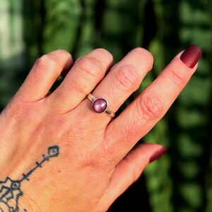 May include: A silver ring featuring a large, oval, pink gemstone. The ring is worn on a hand with dark red nail polish and a small wrist tattoo. The background is a blurred green.