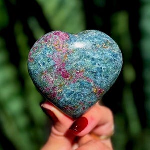 May include: A heart-shaped polished gemstone featuring a blend of deep blue and teal tones with prominent inclusions of vibrant magenta and pink mineral deposits throughout the surface, creating a marbled appearance with a smooth, reflective finish.