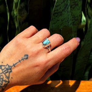 May include: A silver ring featuring a rectangular, turquoise-colored stone. The ring is on a hand with a tattoo on the wrist. The background is a blurred view of green foliage.