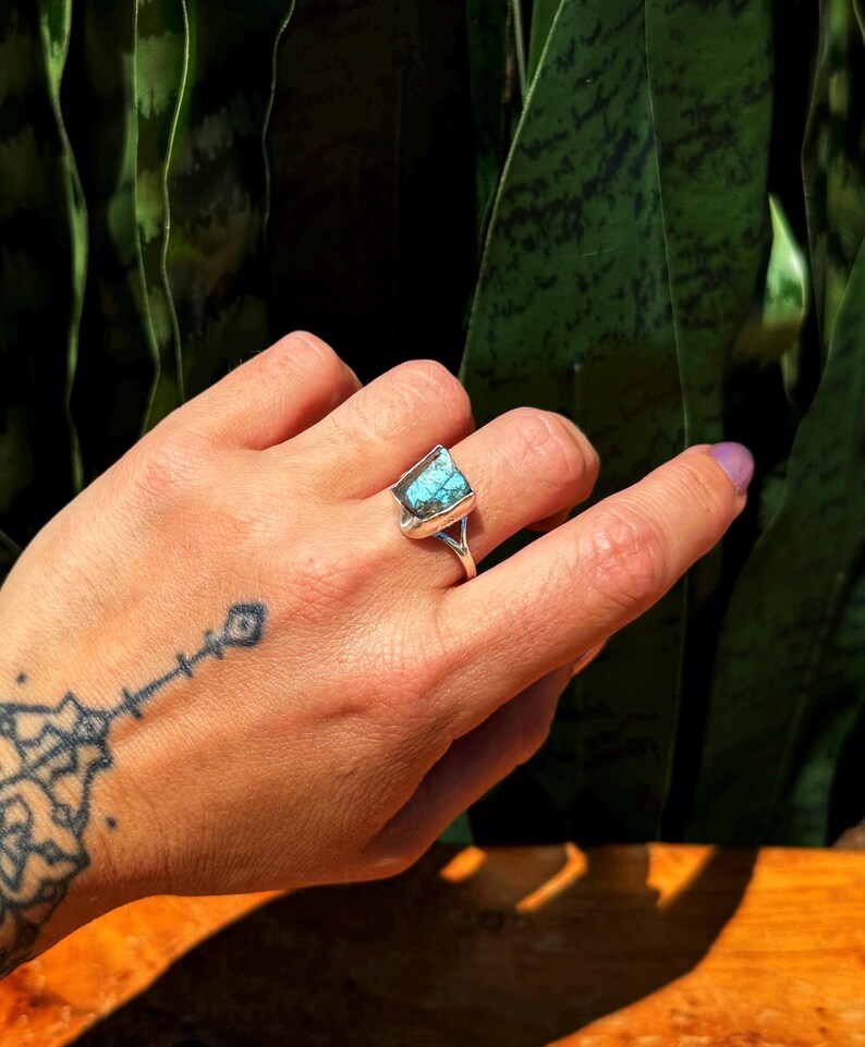 May include: A silver ring featuring a rectangular, turquoise-colored stone. The ring is on a hand with a tattoo on the wrist. The background is a blurred view of green foliage.