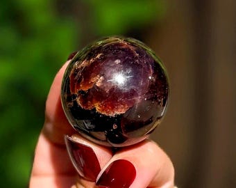 May include: A polished, spherical gemstone with a deep, dark purple and black base color. The surface features prominent, irregular patches of reddish-brown and copper-toned inclusions that catch the light, creating a reflective, glossy appearance.