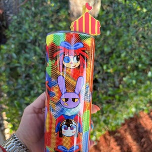 May include: A red, yellow, blue, and green patterned stainless steel tumbler with a cartoon character design. The tumbler has a red and yellow circus tent lid.