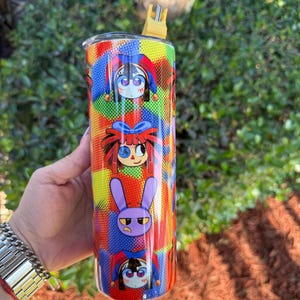 Puede incluir: Un vaso de acero inoxidable colorido con una pajita. El vaso presenta un patr&oacute;n repetitivo de personajes de dibujos animados con diferentes expresiones. Los personajes son un buf&oacute;n, una ni&ntilde;a con el pelo rojo y un conejo morado.