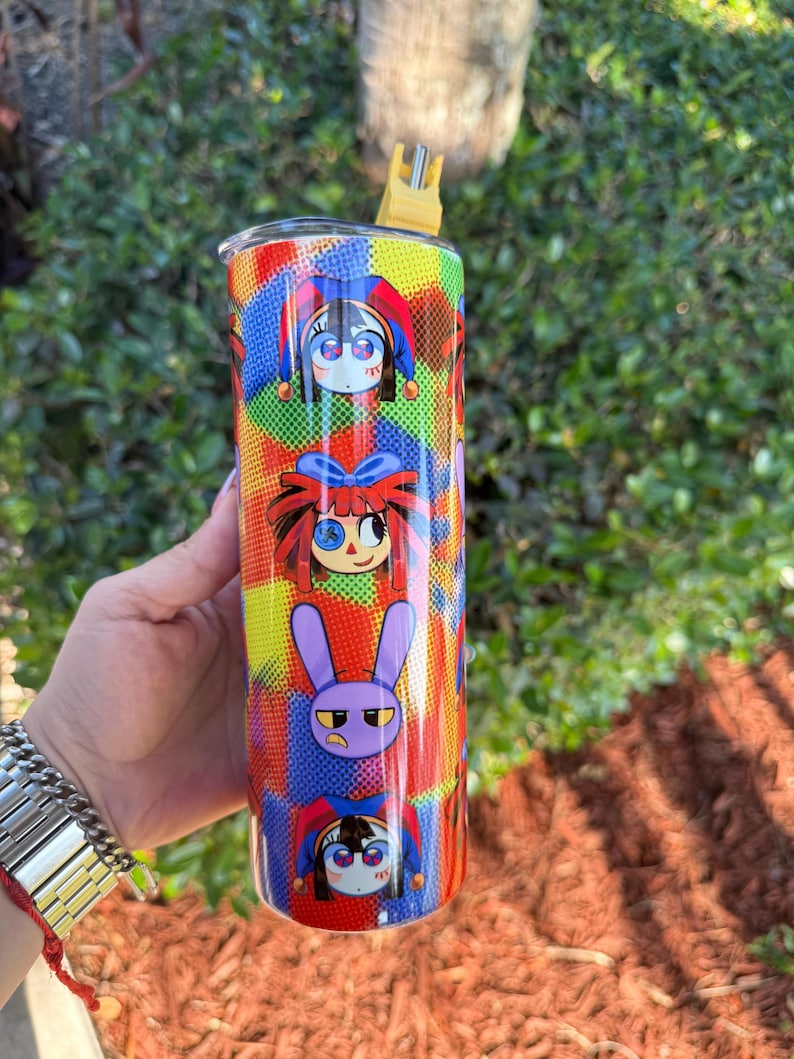 Puede incluir: Un vaso de acero inoxidable colorido con una pajita. El vaso presenta un patr&oacute;n repetitivo de personajes de dibujos animados con diferentes expresiones. Los personajes son un buf&oacute;n, una ni&ntilde;a con el pelo rojo y un conejo morado.
