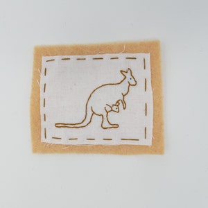 Può includere: Un quadrato di feltro marrone con un quadrato di tessuto bianco all'interno. Il quadrato di tessuto bianco presenta un'immagine ricamata marrone di un canguro con un piccolo nella sua tasca.