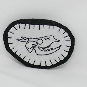 May include: Black and white embroidered patch of a skull with a white background and a black border.