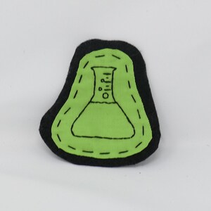 May include: A handmade, black-edged patch featuring a green, embroidered laboratory flask design. The flask has a detailed outline with black stitching, and the patch has a unique, irregular shape. The background is plain white.
