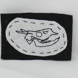 May include: A black felt patch with a white oval embroidered with a black line drawing of a skull. The oval has a white background with a black swirl pattern.
