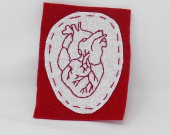 Puede incluir: Un parche de fieltro rojo con un óvalo blanco bordado con un corazón anatómico detallado en hilo rojo.