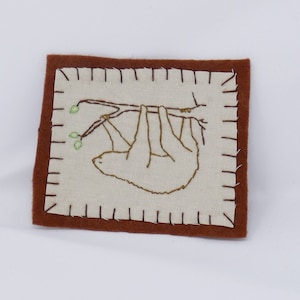 Puede incluir: Un parche rectangular con un borde marrón y un centro color crema. El centro presenta un perezoso bordado colgando de una rama. El perezoso y la rama son marrones, y las hojas son verdes. El parche tiene un borde cosido.