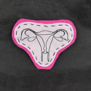 May include: A handmade fabric patch featuring a black embroidered uterus design on white fabric, outlined with a bright pink felt border. The patch is stitched with black thread and has a unique, handcrafted appearance.