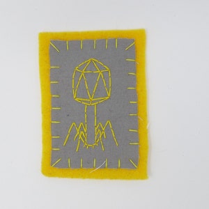 May include: A yellow felt patch with a gray felt center. The center features a yellow embroidered image of a bacteriophage, a type of virus that infects bacteria.