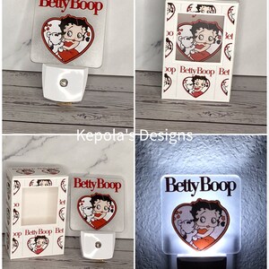 May include: A square, white night light featuring the cartoon character Betty Boop embracing a teddy bear inside a red heart. The words "Betty Boop" are above the heart in red. The night light is plugged into a white outlet.