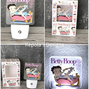 May include: A Betty Boop night light featuring the cartoon character in a pink bathtub with bubbles. The night light has a white base and a square, frosted acrylic panel with the Betty Boop logo and heart designs.
