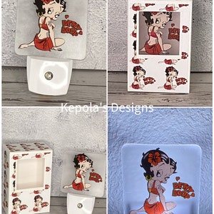 May include: A white night light featuring a square, translucent panel with a Betty Boop graphic. The cartoon character is depicted in a red outfit, with the words "Betty Boop" in red. The night light is plugged into a white base.