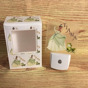 May include: A white night light with a Tiana design. The night light features a princess in a green dress and a frog. The box has a window and repeats the princess and frog design. The word "Tiana" is printed on the night light.