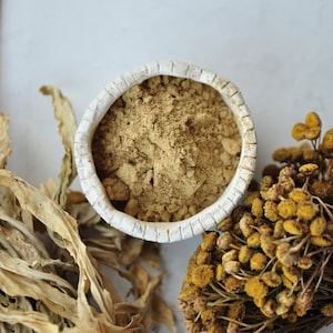 May include: A white ceramic bowl filled with a light brown powder, surrounded by dried herbs. The herbs are a mix of light brown and yellow.