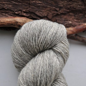 Puede incluir: Primer plano de un ovillo de lana gris. La lana está bien enrollada, mostrando la textura de las fibras. El fondo incluye un trozo de madera oscura y una superficie gris claro.