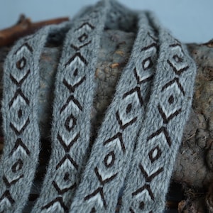 May include: A gray scarf with a geometric diamond pattern in black and white. The scarf is made of a soft, textured material and is draped over a rough, organic surface. The design is repeated along the length of the scarf.
