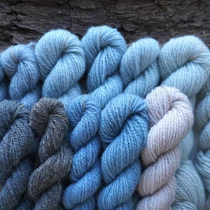 Könnte beinhalten: Eine Sammlung handgefärbter Garnknäuel in verschiedenen Blau-, Grau- und Off-White-Tönen. Das Garn ist in ordentlichen Bündeln angeordnet, die die verschiedenen Texturen und Farben zeigen. Der Hintergrund zeigt Naturholz und Rinde.