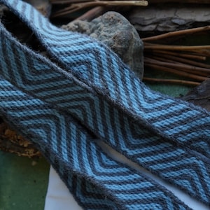 May include: Two woven straps with a chevron pattern in shades of blue and gray. The straps are made of a textured material and appear to be handmade. The background includes natural elements like wood and stone.