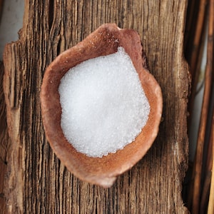 Könnte beinhalten: Eine kleine, braune Keramikschale gefüllt mit weißem, granuliertem Zucker. Die Schale steht auf einem Stück verwittertem, braunem Holz.