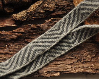 May include: A woven fabric ribbon featuring a repeating chevron pattern in shades of charcoal grey and light grey. The textile has a textured, fibrous appearance with visible yarn strands running diagonally across the surface of the material.
