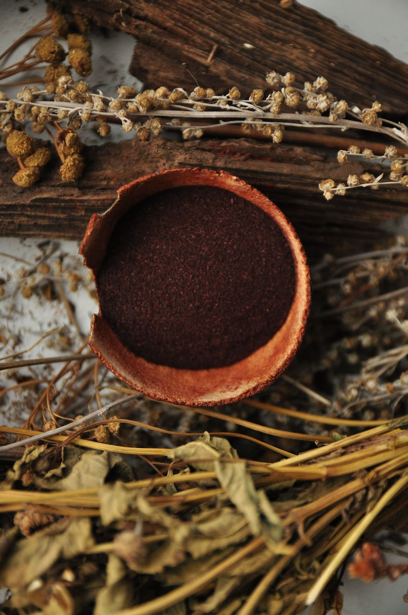 Op de afbeelding: Een kleine, ronde terracotta kom gevuld met een donkerrood poeder. De kom is omringd door gedroogde, bruine plantenstengels en bladeren.