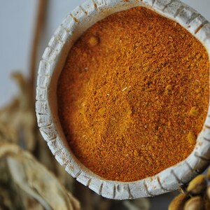 Pu&ograve; includere: Una piccola ciotola di ceramica bianca piena di polvere di spezie di colore arancione brillante. La ciotola ha un bordo decorativo e si trova su una superficie di colore marrone chiaro.