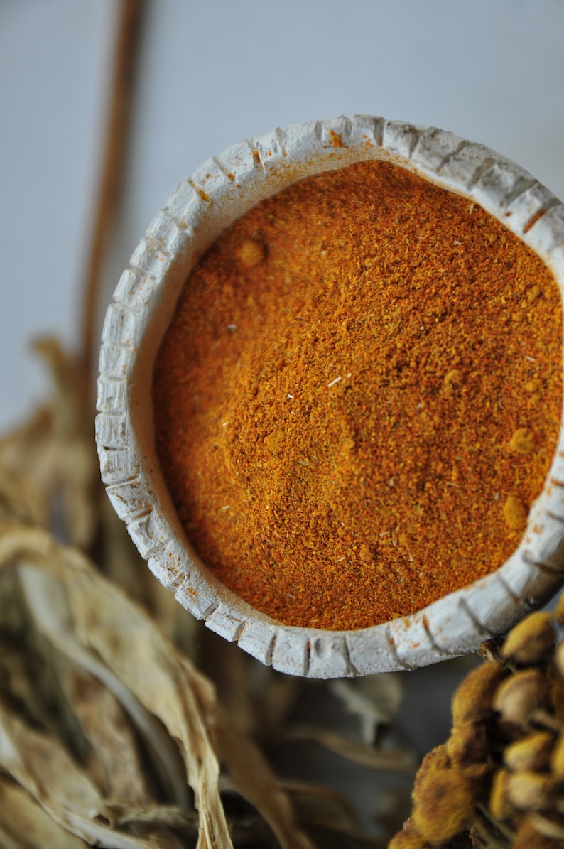 Pu&ograve; includere: Una piccola ciotola di ceramica bianca piena di polvere di spezie di colore arancione brillante. La ciotola ha un bordo decorativo e si trova su una superficie di colore marrone chiaro.