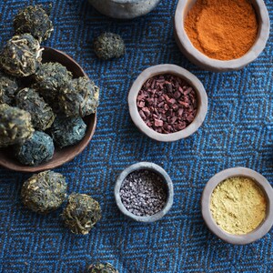 Op de afbeelding: Een verzameling natuurlijke kleurstoffen in kleine kommen op een blauw-zwarte, gepatroneerde stof. De kleurstoffen omvatten indigo, paprika, rode wortel, zwart zout en kurkuma.