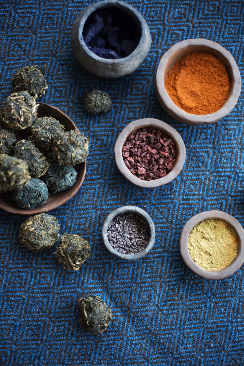Op de afbeelding: Een verzameling natuurlijke kleurstoffen in kleine kommen op een blauw-zwarte, gepatroneerde stof. De kleurstoffen omvatten indigo, paprika, rode wortel, zwart zout en kurkuma.