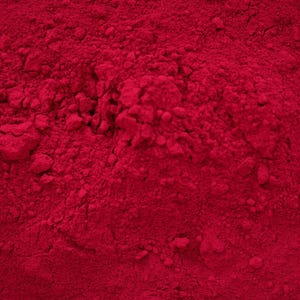 May include: A fine, vibrant magenta-red powder with a granular, uneven texture. The surface displays small clumps and subtle variations in depth, creating a dense, monochromatic field of intense, saturated pigment across the entire frame.