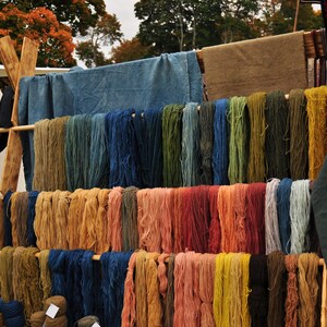 Op de afbeelding: Een houten rek met verschillende kleuren garen dat eraan hangt. Het garen is in rijen gerangschikt en heeft verschillende lengtes. Het garen is in verschillende kleuren, waaronder blauw, groen, rood, oranje en geel.