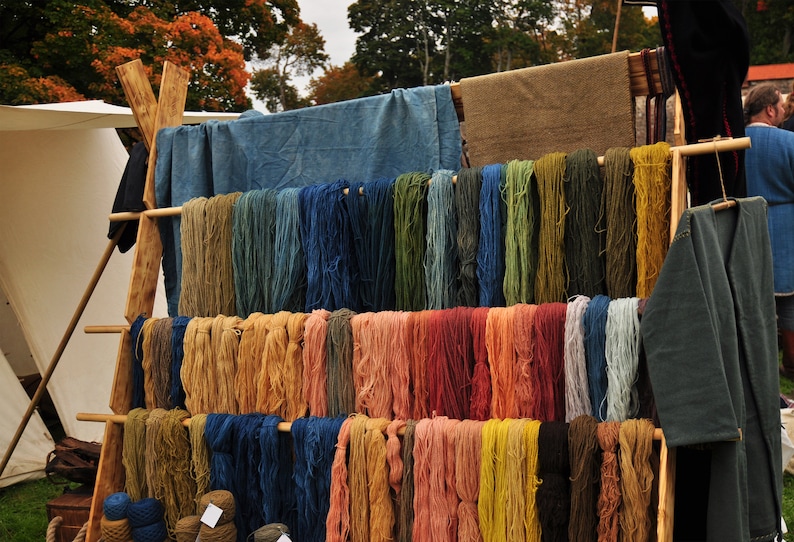 Op de afbeelding: Een houten rek met verschillende kleuren garen dat eraan hangt. Het garen is in rijen gerangschikt en heeft verschillende lengtes. Het garen is in verschillende kleuren, waaronder blauw, groen, rood, oranje en geel.