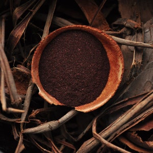 Op de afbeelding: Een kleine, ronde terracotta kom gevuld met een donkerrood poeder, omringd door bruin, gedroogd plantenmateriaal.