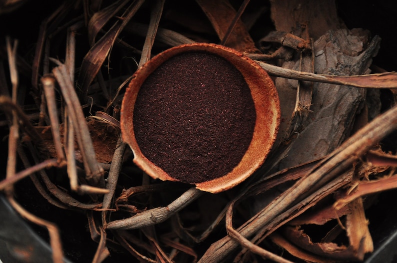Op de afbeelding: Een kleine, ronde terracotta kom gevuld met een donkerrood poeder, omringd door bruin, gedroogd plantenmateriaal.