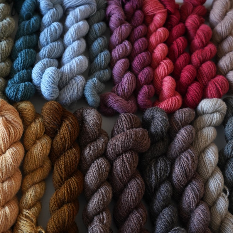 May include: A collection of yarn skeins in various shades of blue, purple, red, brown, and gray. The yarn is arranged in rows, with each row featuring a different color.