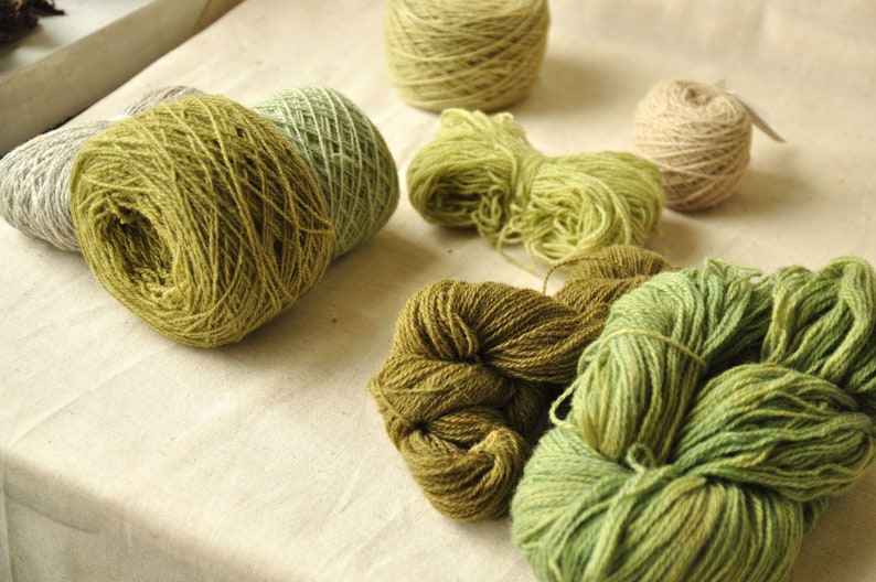 May include: Assortment of yarn skeins in various shades of green and beige. The yarn is arranged on a white surface.