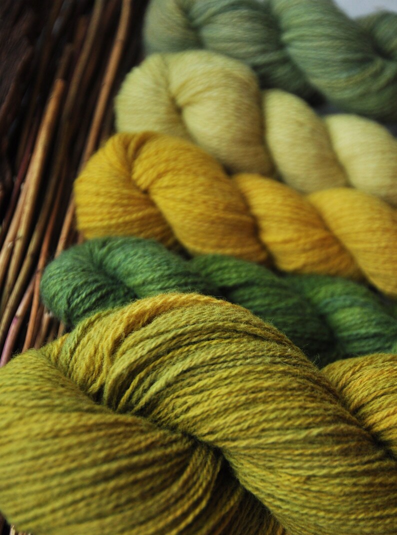 May include: A collection of yarn skeins in shades of green and yellow. The yarn is arranged in a stack, with the darkest green skein at the bottom and the lightest yellow skein at the top.