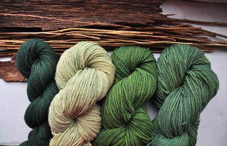 May include: Four skeins of yarn in shades of green and beige. The yarn is arranged in a figure of eight shape.