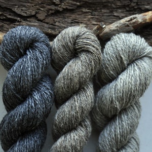 May include: Three skeins of yarn in shades of grey, black, and white. The yarn is twisted and piled on top of a piece of driftwood.