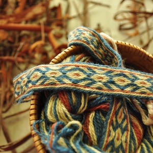 May include: A woven tapestry with blue, white and red stripes in a basket. The tapestry is made of yarn and has a geometric pattern.