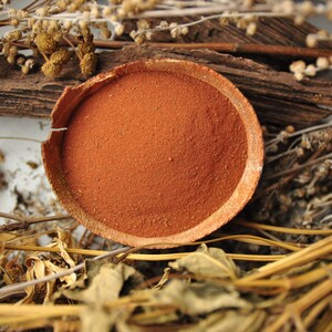 Puede incluir: Un cuenco redondo de color terracota lleno de polvo fino de color marr&oacute;n rojizo. El cuenco est&aacute; rodeado de plantas secas, ramitas y trozos de madera, creando una est&eacute;tica natural y terrosa. El borde del cuenco est&aacute; ligeramente agrietado.