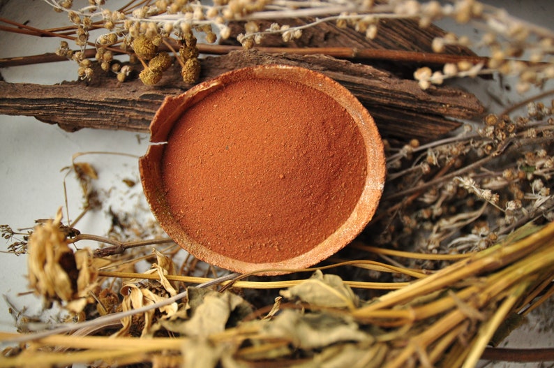 Puede incluir: Un cuenco redondo de color terracota lleno de polvo fino de color marr&oacute;n rojizo. El cuenco est&aacute; rodeado de plantas secas, ramitas y trozos de madera, creando una est&eacute;tica natural y terrosa. El borde del cuenco est&aacute; ligeramente agrietado.