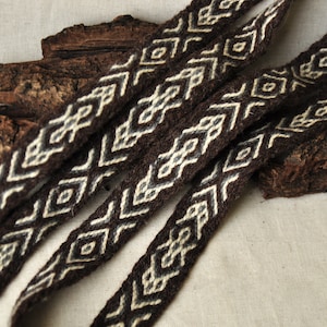May include: Close-up of brown woven fabric trim with a white geometric pattern. The trim is arranged in a criss-cross pattern on a light brown surface.