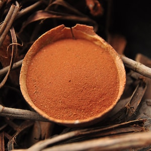 May include: A wooden bowl filled with reddish-brown powder, set amongst dried twigs and leaves. The powder has a fine texture. The natural arrangement suggests a rustic or organic theme, with the bowl's contents appearing finely ground.