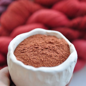 Op de afbeelding: Een witte keramische kom gevuld met een roodbruin poeder, met een wazige achtergrond van rood garen.