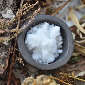 May include: A close-up of a small, dark gray ceramic bowl filled with white, fluffy crystals. The bowl is sitting on a rustic background of dried plants and bark.